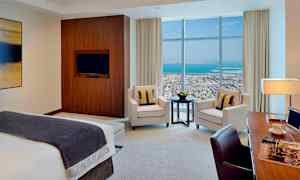 Moderni hotellihuone, jossa sänky, nojatuolit, pöytä ja työtila. Suuret ikkunat tarjoavat näkymän kaupunkiin ja merelle. (Tekoälyn luoma teksti)