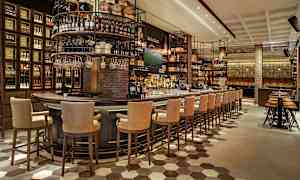 Kuva modernin baarin sisätiloista, jossa on korkeat selkänojatuolit suuren baaritiskin äärellä, ympärillä hyllyjä, jotka ovat täynnä pulloja ja tynnyreitä. (Tekoälyn luoma teksti)