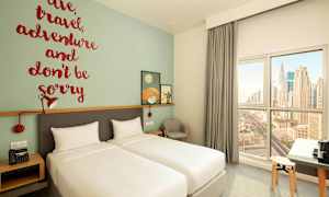 Hotellihuone, jossa on kaksi erillistä vuodetta, seinäkoriste inspiroivalla lainauksella ja ikkuna näkymällä kaupungin siluettiin. (Tekoälyn luoma teksti)