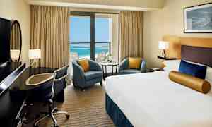 Hotellihuone, jossa suuri sänky, työpöytä ja näkymä siniselle merelle parvekkeen lasiovien läpi. (Tekoälyn luoma teksti)