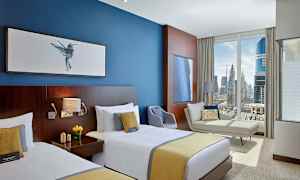 Hotellihuone, jossa on kaksi yhden hengen vuodetta, sininen seinä tauluineen, sohva, nojatuoli ja suuri ikkuna, josta avautuu näkymä kaupunkiin. (Tekoälyn luoma teksti)