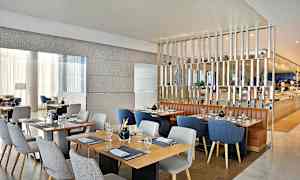 Kuva modernista ravintolasta, jossa on useita katettuja pöytiä ja erivärisiä tuoleja. Lämmin ja tyylikäs sisustus. (Tekoälyn luoma teksti)