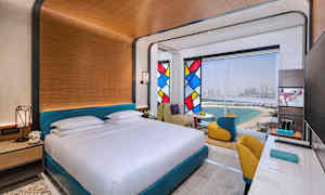 Hotellihuone, jossa on suuri sänky, värikkäitä lasimaalauksia ja näkymä rannalle ja kaupunkiin. (Tekoälyn luoma teksti)