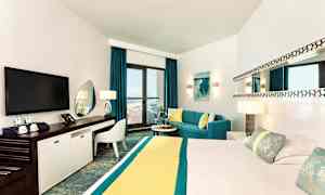 Moderni hotellihuone, jossa on suuri sänky, televisio, työtila ja oleskelualue, josta on pääsy parvekkeelle merinäköalalla. (Tekoälyn luoma teksti)