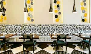 Ravintolasisustus, jossa mustavalkoiset lattialaatat, kuvioidut seinäpaneelit sekä koristeellinen istuinpenkki mustien tuolien ympäröimin pöydin. Värikkäitä seinäkoristeita näkyy seinällä. (Tekoälyn luoma teksti)