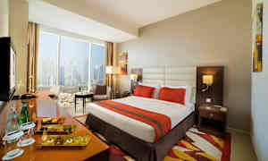 Hotellihuone, jossa suuri sänky, oranssi peite, ikkuna kaupunkinäkymällä ja istuinryhmä. (Tekoälyn luoma teksti)