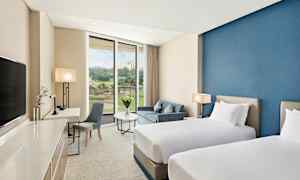 Hotellihuone, jossa on kaksi erillissänkyä, sininen seinä, istuinryhmä pöydällä ja sohvalla sekä suuri ikkuna, josta näkyy maisema. (Tekoälyn luoma teksti)