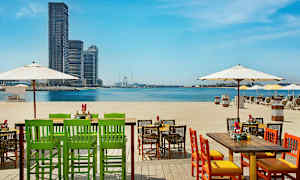 Tuolit ja pöydät rannalla, näkymä merelle ja kaupunkinäkymä etäällä. (Tekoälyn luoma teksti)