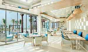 Valoisa ja moderni ravintola, jossa tuoleissa siniset tyynyt ja näkymä altaalle ja palmuihin. (Tekoälyn luoma teksti)