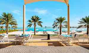 Ulkoterassi, jossa on pöytiä, tuoleja ja näkymä trooppisiin palmuihin, valkoiselle hiekkarannalle ja merelle. (Tekoälyn luoma teksti)