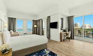 Moderni hotellihuone, jossa on iso sänky, työpöytä, parveke ja näkymä siniselle taivaalle ja rakennuksille. (Tekoälyn luoma teksti)