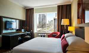 Kuva modernista hotellihuoneesta, jossa on sänky etualalla, työpöytä television kanssa ja ikkuna kaupunkinäkymällä. (Tekoälyn luoma teksti)