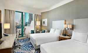 Hotellihuone, jossa on kaksi parisänkyä, kirjoituspöytä tuolilla ja parveke näkymällä moderneihin rakennuksiin. (Tekoälyn luoma teksti)