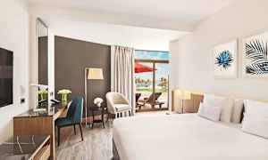 Hotellihuone, jossa on modernit kalusteet, suuri sänky, marmoripöytä ja terassi, jossa on aurinkotuoleja ja näkymä palmupuihin. (Tekoälyn luoma teksti)