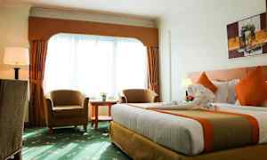 Hotellihuone, jossa on suuri sänky, kaksi nojatuolia, pyöreä pöytä ja suuret verhoilla varustetut ikkunat. (Tekoälyn luoma teksti)