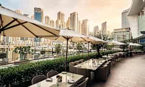 Ulkoilmaravintola, jossa on aurinkovarjoja ja pöytiä, sataman äärellä ja taustalla korkeat rakennukset. (Tekoälyn luoma teksti)