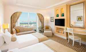 Hotellihuone, jossa on kaksi yhden hengen sänkyä, sohva lähellä ikkunaa näkymällä, tuoli ja pöytä peilillä. (Tekoälyn luoma teksti)