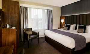 Hotellihuone, jossa on suuri sänky, nojatuoli ja puisia yksityiskohtia. (Tekoälyn luoma teksti)