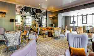 Moderni hotellihuone mukavilla istuimilla, tyylikkäillä koristeilla ja seinätapetilla, jossa on tekstiä ja taidetta. Suuret ikkunat päästävät luonnonvalon sisään. (Tekoälyn luoma teksti)