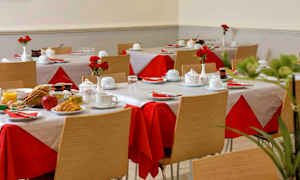 Huone, jossa on katettuja pöytiä, punaiset ja valkoiset pöytäliinat, sekä aamiaisruokaa lautasilla. (Tekoälyn luoma teksti)