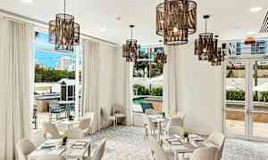 Valoisa ja tyylikkäästi sisustettu huone, jossa on valkoiset huonekalut, suuret ikkunat ja riippuvalaisimet. Näkymässä näkyy uima-allas ja ympäröivät rakennukset. (Tekoälyn luoma teksti)