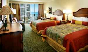 Hotellihuone, jossa on kaksi vuodetta värikkäillä päiväpeitteillä, kirjoituspöytä ja avoin parveke merinäköalalla. (Tekoälyn luoma teksti)