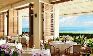 Ravintola, jonka pöydät ovat katettu valkoisilla pöytäliinoilla ja kukilla. Taustalla näkyy merinäkymä ja sininen taivas. (Tekoälyn luoma teksti)