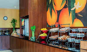 Buffet-pöytä, jossa on leipää, hedelmiä ja pieniä tarjoilukulhoja, värikkään hedelmämaalauksen edessä. (Tekoälyn luoma teksti)