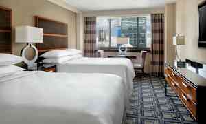 Hotellihuone, jossa on kaksi parisänkyä valkoisilla vuodevaatteilla, pöytä ja tuolit ikkunan lähellä sekä lipasto, jonka päällä on televisio. (Tekoälyn luoma teksti)