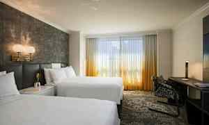 Hotellihuone, jossa on kaksi suurta vuodetta, kirjoituspöytä ja suuret ikkunat, joissa on päivänvaloa läpäisevät verhot. (Tekoälyn luoma teksti)