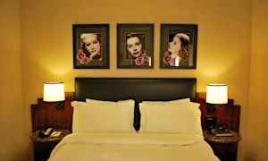 Hotellihuoneen sisustus, jossa on parivuode, kaksi yöpöytää lampuilla ja maalauksia sängyn yläpuolella. (Tekoälyn luoma teksti)