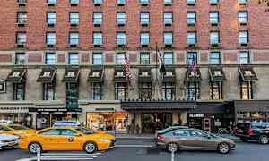 Tiilirakennus, jonka sisäänkäyntien yllä on markiisit, liikennettä autoilla ja takseilla kadulla. (Tekoälyn luoma teksti)