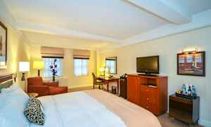 Hotellihuone, jossa on suuri sänky, nojatuoli, kirjoituspöytä ja televisio lipaston päällä. (Tekoälyn luoma teksti)