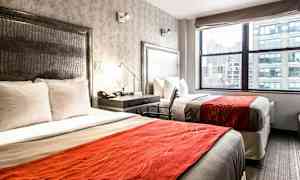 Hotellihuone, jossa on kaksi vuodetta valkoisilla vuodevaatteilla ja oransseilla peitteillä. Ikkunasta on näkymä kaupunkiin. (Tekoälyn luoma teksti)