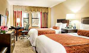 Hotellihuone, jossa on kaksi erillistä vuodetta, kirjoituspöytä tuoleineen, lamppu ja ikkunat kaupunkiin päin. (Tekoälyn luoma teksti)