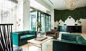 Moderni hotellin aulatila, jossa on sohvia, nojatuoleja ja pöytä etualalla. Taustalla näkyy vastaanotto, jossa on kyltti ja vihreä kasviseinä. (Tekoälyn luoma teksti)