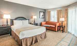 Hotellihuone, jossa on parivuode, yöpöydät kummallakin puolella, pieni sohva, sohvapöytä ja kolme lamppua. Ikkunat verhoineen näkyvät taustalla. (Tekoälyn luoma teksti)