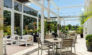 Ulkoterassi, jossa on valkoiset huonekalut ja vihreitä kasveja, pergolarakenteen alla. (Tekoälyn luoma teksti)