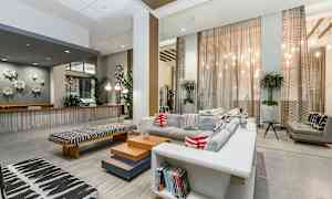 Moderni olohuone, jossa on suuret ikkunat, harmaa sohva värikkäillä tyynyillä sekä sisustuselementtejä, kuten ruukkukasveja ja tietokirjoja. (Tekoälyn luoma teksti)