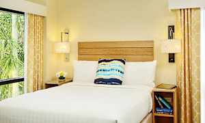 Makuuhuone, jossa sänky valkoisilla vuodevaatteilla, koristeellinen tyyny ja yöpöytä. Keltaisella kuviolla varustetut verhot kehystävät ikkunat. (Tekoälyn luoma teksti)