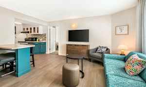 Olohuone, jossa sohva, nojatuoli ja televisio seinällä. Keittiöosa sinisillä kaapeilla näkyy vasemmalla. (Tekoälyn luoma teksti)