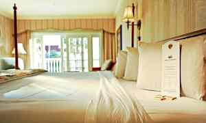 Vuode hotellihuoneessa, jossa on valkoiset lakanat ja tyynyt, sekä parvekkeen oven kautta sisään tuleva valo. (Tekoälyn luoma teksti)