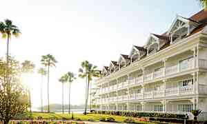 Valkoinen hotelli, jossa on useita kerroksia ja parvekkeellisia sisäänkäyntejä, ympärillä palmuja ja kukkien koristama nurmikko. (Tekoälyn luoma teksti)