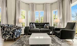 Moderni olohuone, jossa on sohva, kaksi nojatuolia, keskellä sohvapöytä ja suuret ikkunat, jotka päästävät luonnonvaloa sisään. (Tekoälyn luoma teksti)