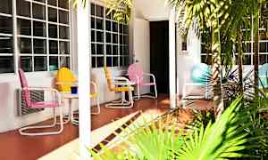 Värikkäitä metallituoleja ja pöytä sijaitsevat terassilla talon edessä, jossa on suuret ikkunat. Terassin edessä on vihreitä palmuja. (Tekoälyn luoma teksti)