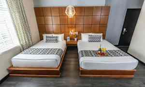 Kuva hotellihuoneesta, jossa on kaksi erillistä sänkyä valkoisilla vuodevaatteilla ja kuvioiduin päiväpeittein. Huoneessa on puupaneeliseinä ja sänkyjen välissä yöpöytä, jolla on kukka. (Tekoälyn luoma teksti)