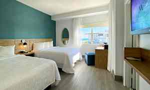 Moderni hotellihuone, jossa on kaksi parisänkyä, työpöytä ja suuri ikkuna, josta pääsee luonnonvaloa sisään. (Tekoälyn luoma teksti)