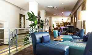 Kuva hotellin aulasta, jossa on sinisiä istuimia, vihreitä kasveja ja baari taustalla. (Tekoälyn luoma teksti)