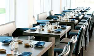 Tyylikäs ravintola-alue, jossa katettuja pöytiä, siniset lautasliinat ja lasit. Taustalla vaaleat ikkunat. (Tekoälyn luoma teksti)