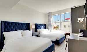 Hotellihuone, jossa on kaksi sänkyä sinisillä sängynpäädyillä, yöpöytä lampun kanssa sänkyjen välissä ja työpöytä, jossa on kukka ja kahvikone. (Tekoälyn luoma teksti)